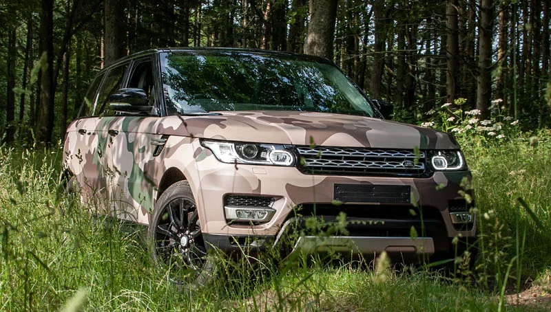 A camouflage-covered Land Rover parked among trees in a dense forest setting.
