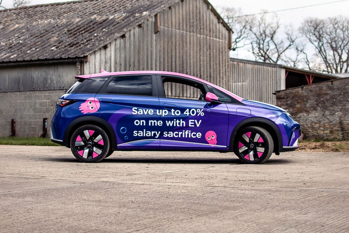 A blue electric hatchback parked outdoors features a vibrant purple promotional vehicle wrap with the message "Save up to 40% on me with EV salary sacrifice" and pink cartoon graphics.