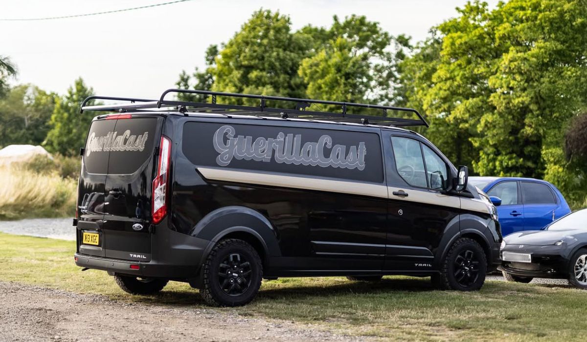 A black van parked along the side of a road, surrounded by greenery and a clear blue sky.