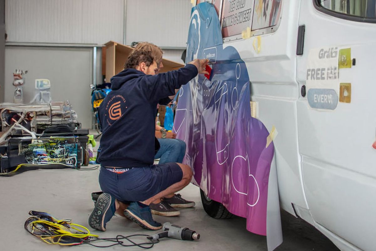 A technician kneels beside a white van, carefully applying a large blue and purple vinyl wrap with a squeegee inside a vehicle workshop. A heat gun and installation tools lie on the floor nearby.